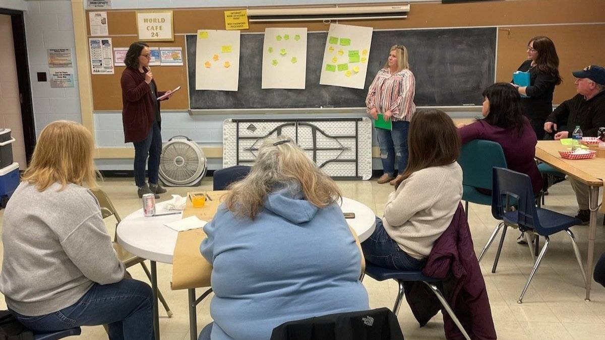 A photo of the World Cafe event with Michelle Miller holding a microphone and speaking to an audience in a classroom